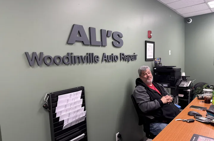 John smiling behind the front desk at Ali's Woodinville Auto Repair, with the shop's name on the wall behind him