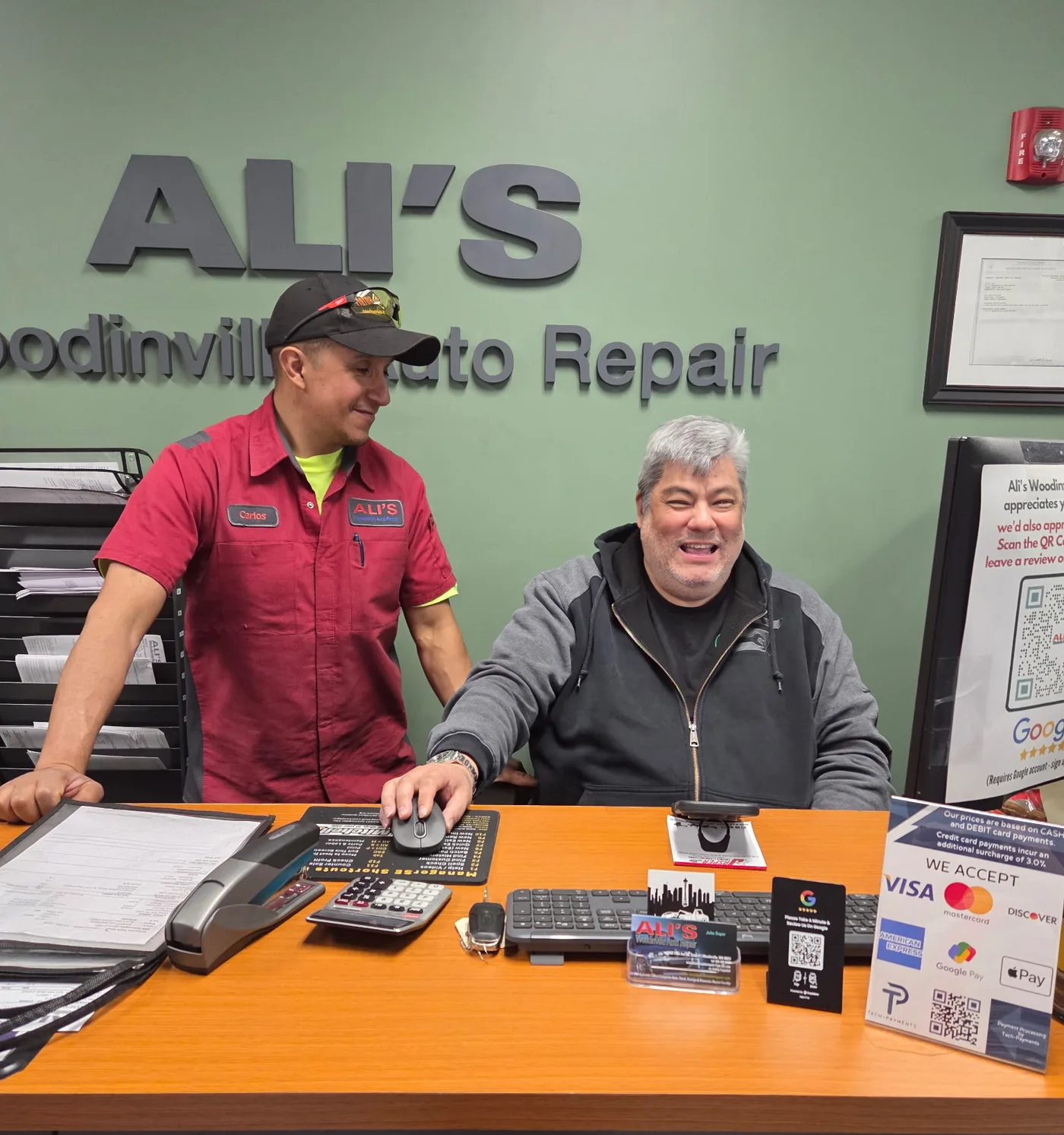 Carlos and John at the front desk of Ali's Woodinville Auto Repair, under the shop's name on the green office wall