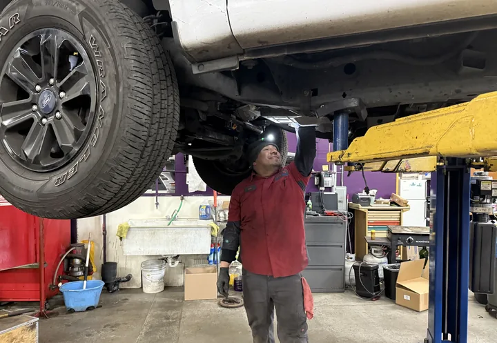 A technician at Ali's Woodinville Auto Repair inspecting the underside of a pickup truck on the lift with a flashlight
